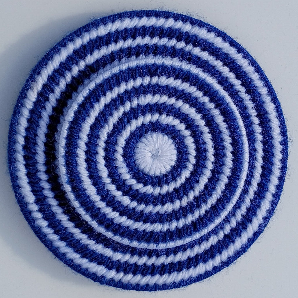 A round, hand-sewn playing card holder made from yarn and plastic canvas with concentric spiral stripes in dark blue and white, photographed from directly above on a light gray background.