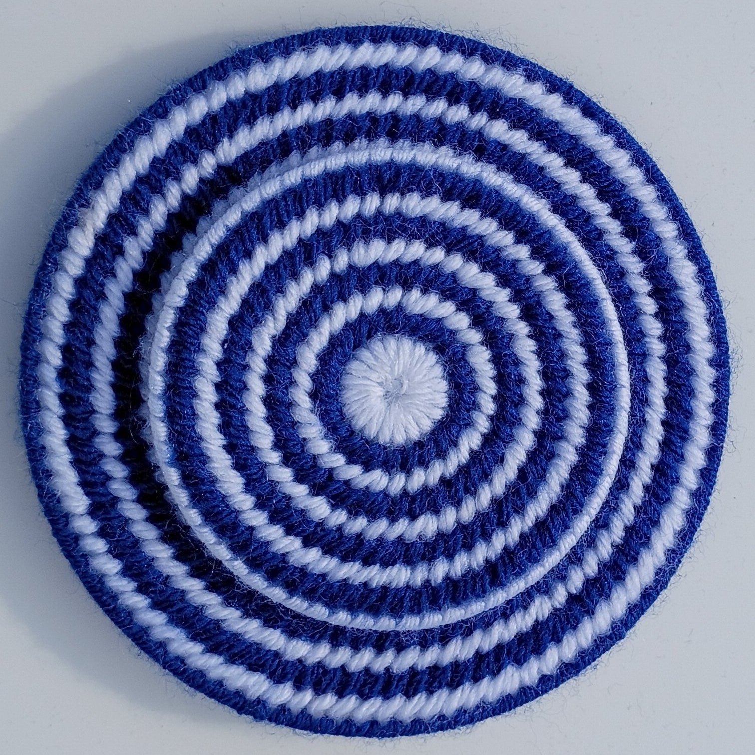 A round, hand-sewn playing card holder made from yarn and plastic canvas with concentric spiral stripes in dark blue and white, photographed from directly above on a light gray background.